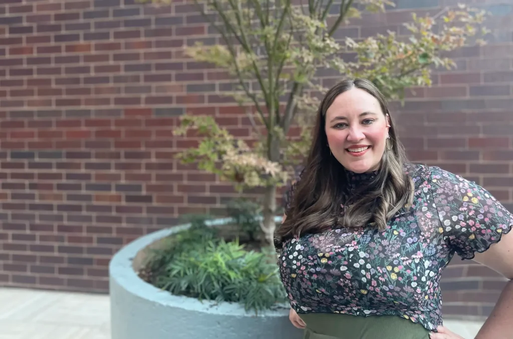 A picture of Taylor Desens. Taylor stand in front of a brick wall, a tree between them. She stands hands akimbo and is wearing a flower-printed dark shirt and green pants.