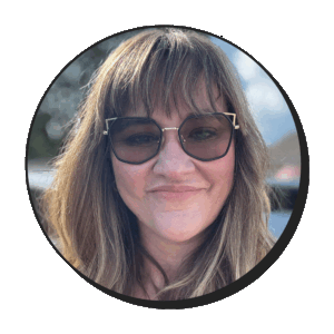 A woman with long brown hair and bangs, wearing large sunglasses, smiling at the camera. The photo is in a circular frame with a blurred outdoor background.