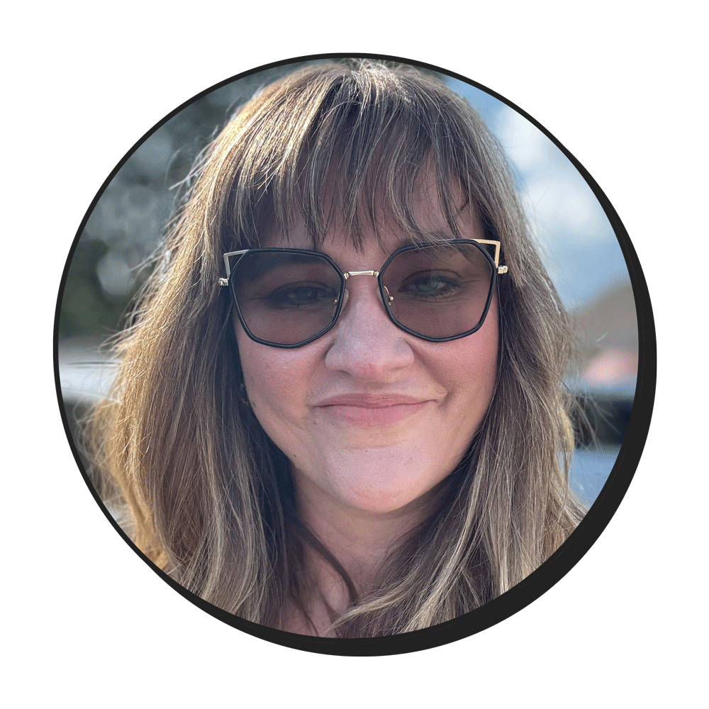 A woman with long brown hair and bangs, wearing large sunglasses, smiling at the camera. The photo is in a circular frame with a blurred outdoor background.