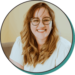 A woman with long, wavy light brown hair and glasses is smiling at the camera, wearing a white top and sitting indoors against a light background.