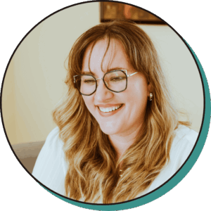 A woman with long, wavy blonde hair and glasses is smiling while looking slightly downward, wearing a white top.