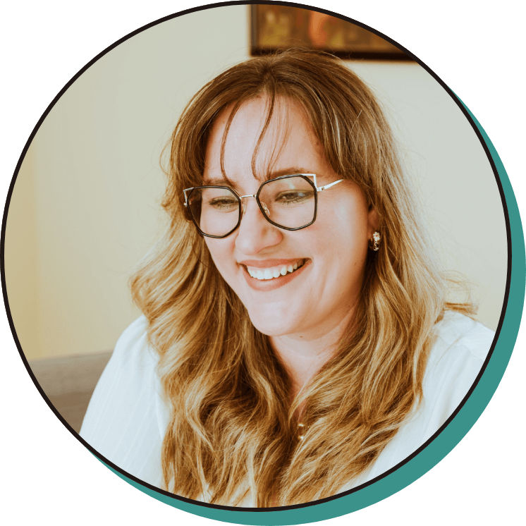 A woman with long, wavy blonde hair and glasses is smiling while looking slightly downward, wearing a white top.