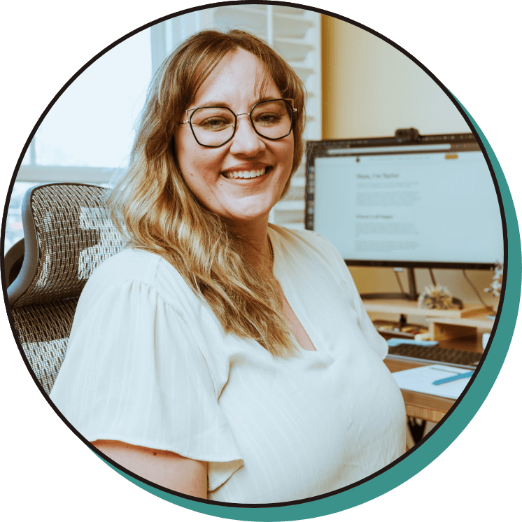 A woman with glasses and a white blouse sits smiling at her desk in front of a computer monitor displaying a document.