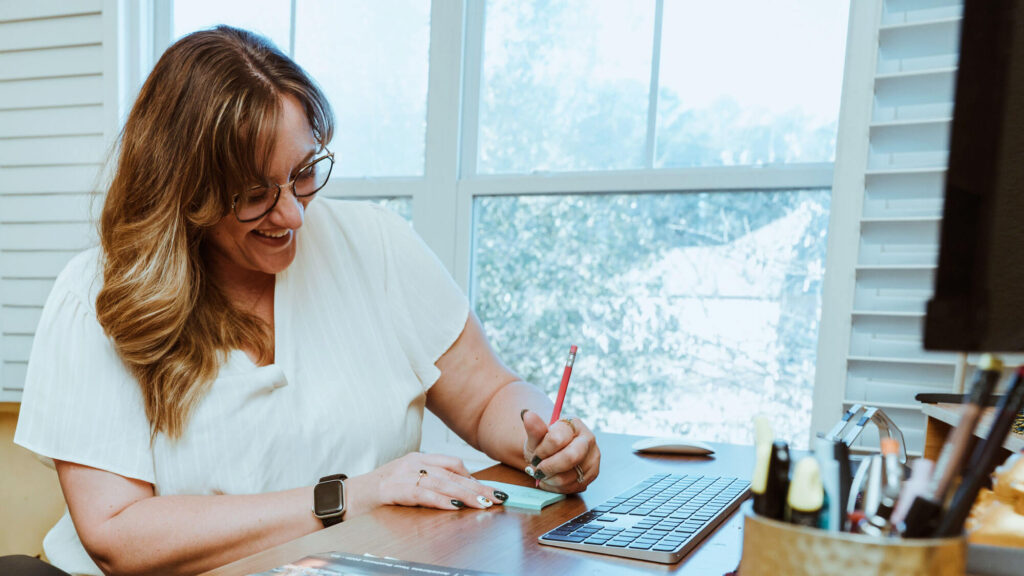 A woman sits at a desk, smiling and writing on a notepad with a pencil, next to a keyboard and office supplies, with windows in the background.