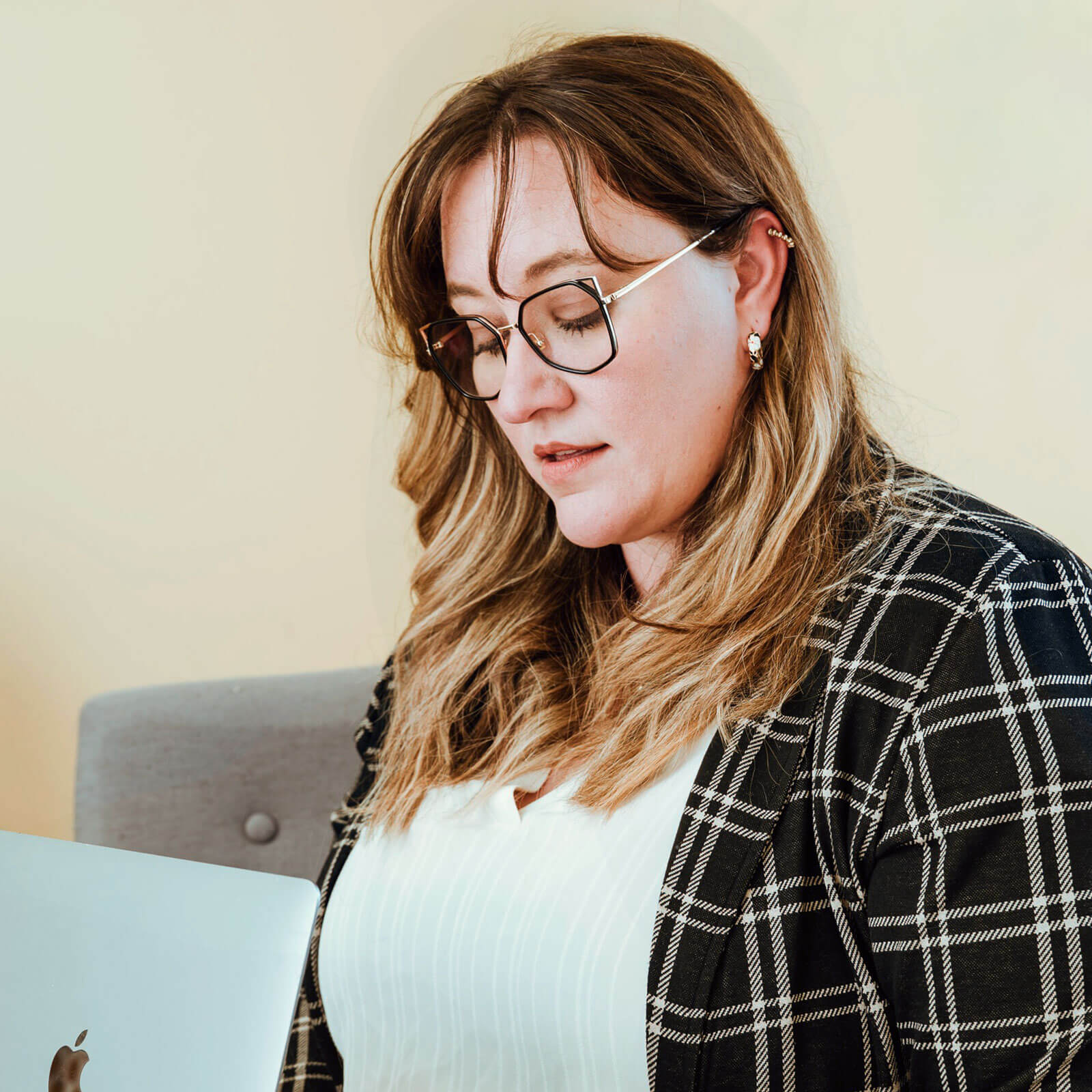 A woman with glasses and long hair, wearing a plaid jacket and white shirt, looks down while seated near a silver laptop.