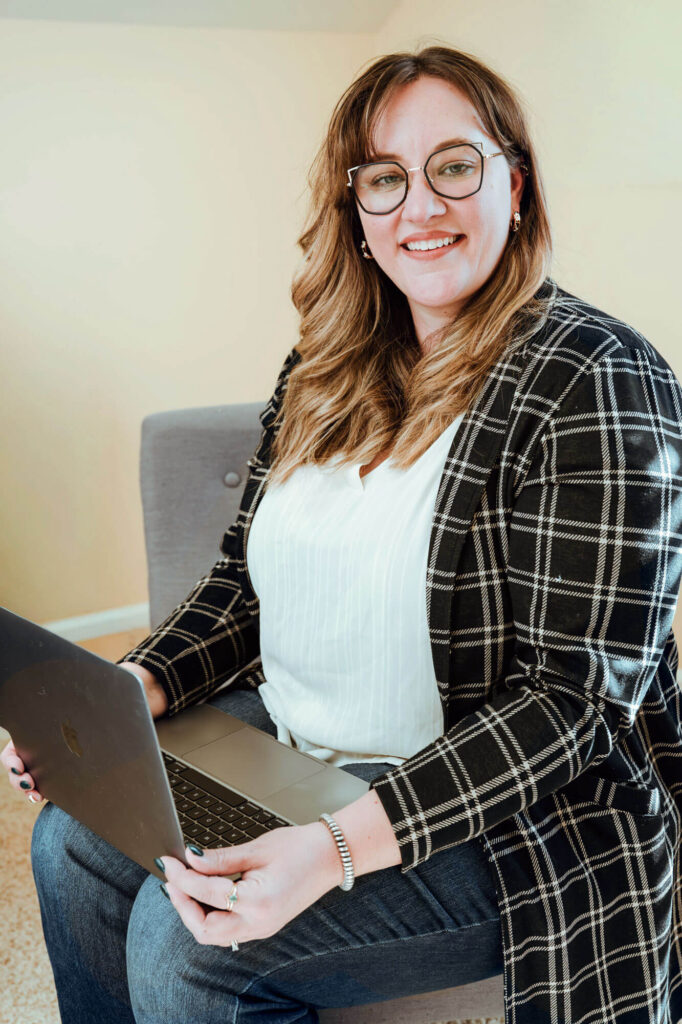 A woman with glasses and long hair sits on a gray chair, smiling at the camera while holding an open laptop on her lap.