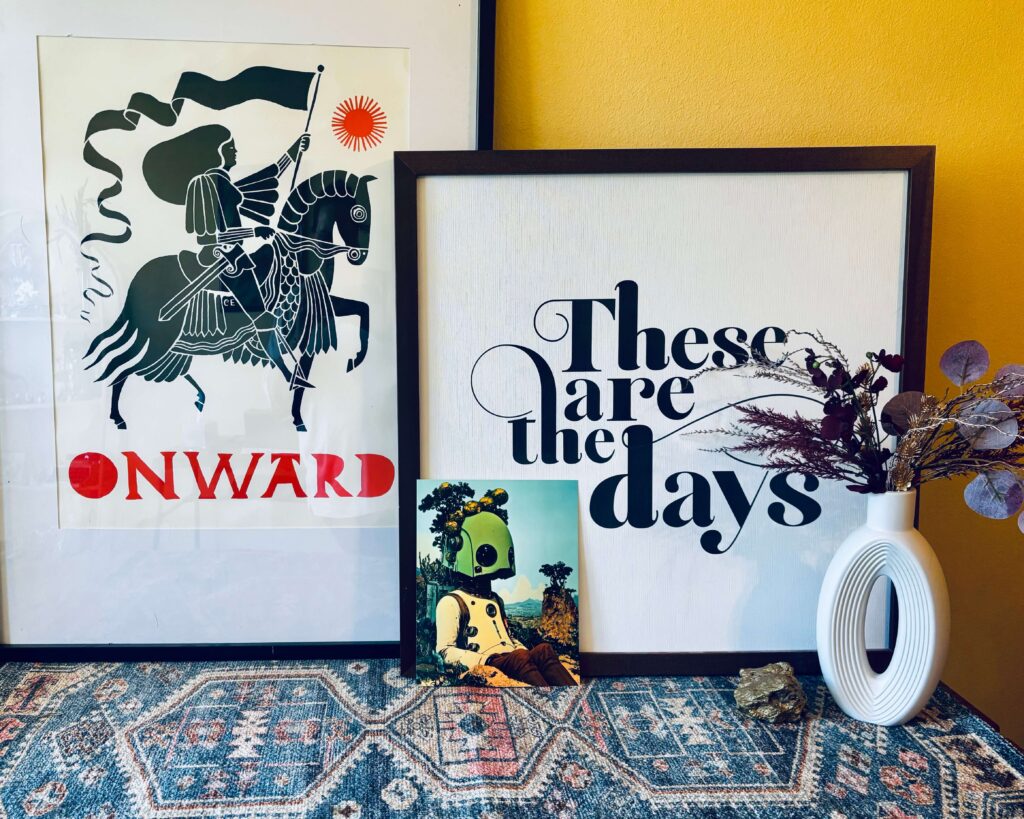 Framed Onward knight print, Taylor “These are the days” sign, small robot photo, and white vase with dried flowers arranged on a patterned rug against a yellow wall.