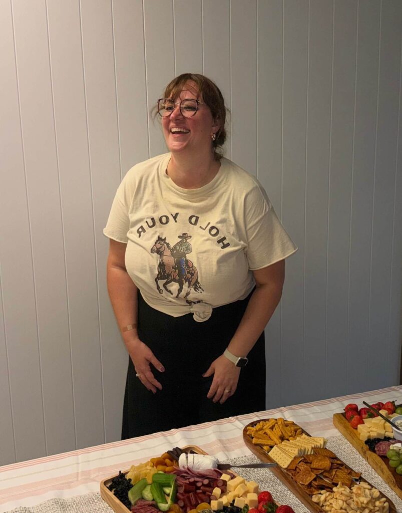 A woman wearing glasses and a "Hold Your Horses" t-shirt stands smiling by a snack-filled table with cheese, fruit, and crackers set against a light-paneled wall.